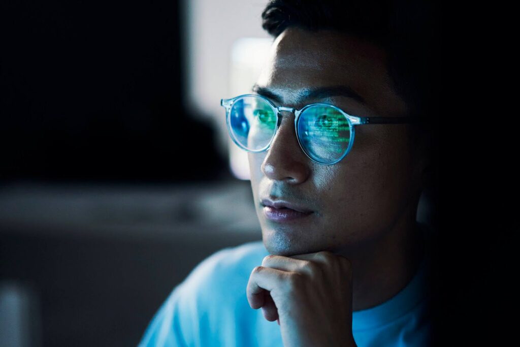 A computer science student stares at a computer screen; his eyeglass lenses reflect the screen text.