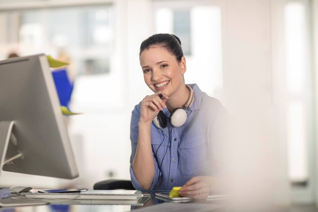 A woman with noise-canceling headphones looks up from her desktop computer to smile at the camera.