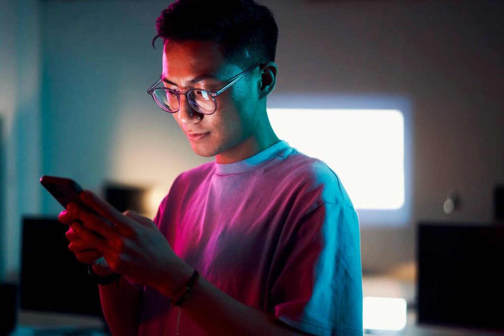 Man in a tech office checks his cell phone; multiple computer monitors are visible in the background.