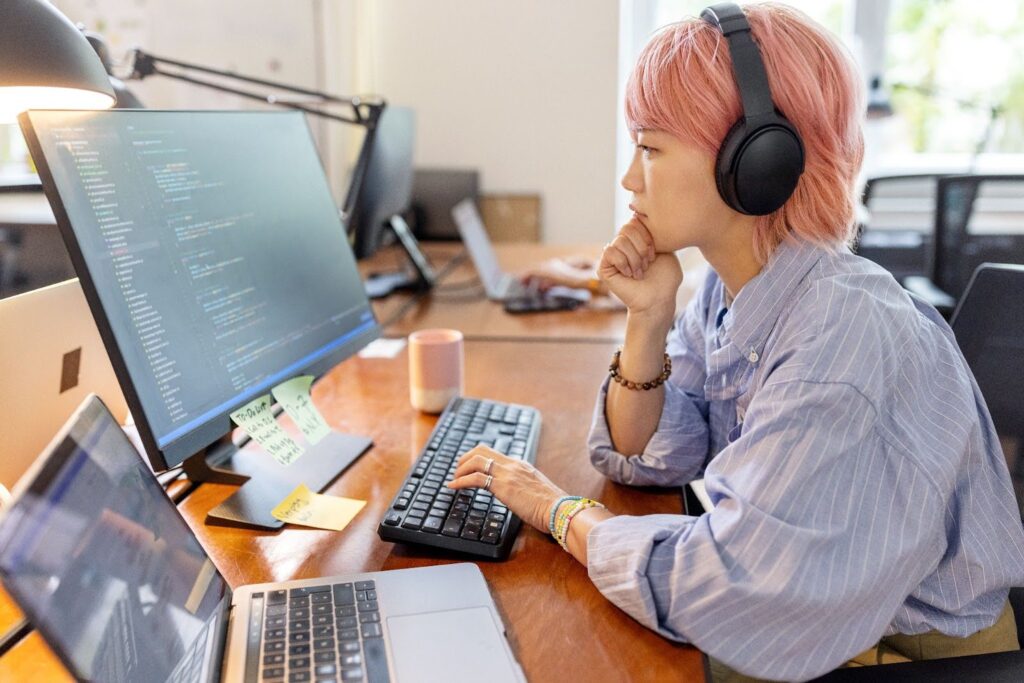 A developer sits at a computer station with multiple computers; coding script is visible on her external monitor.