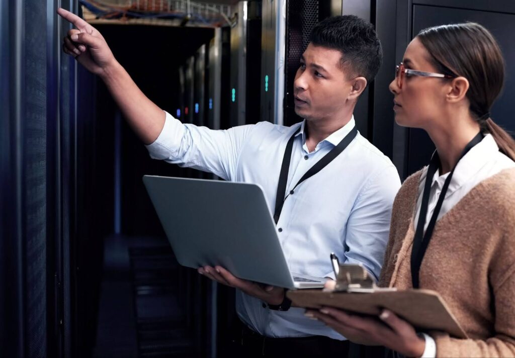 Two technicians work together in a server room; one holds a laptop, the other a clipboard and pen.