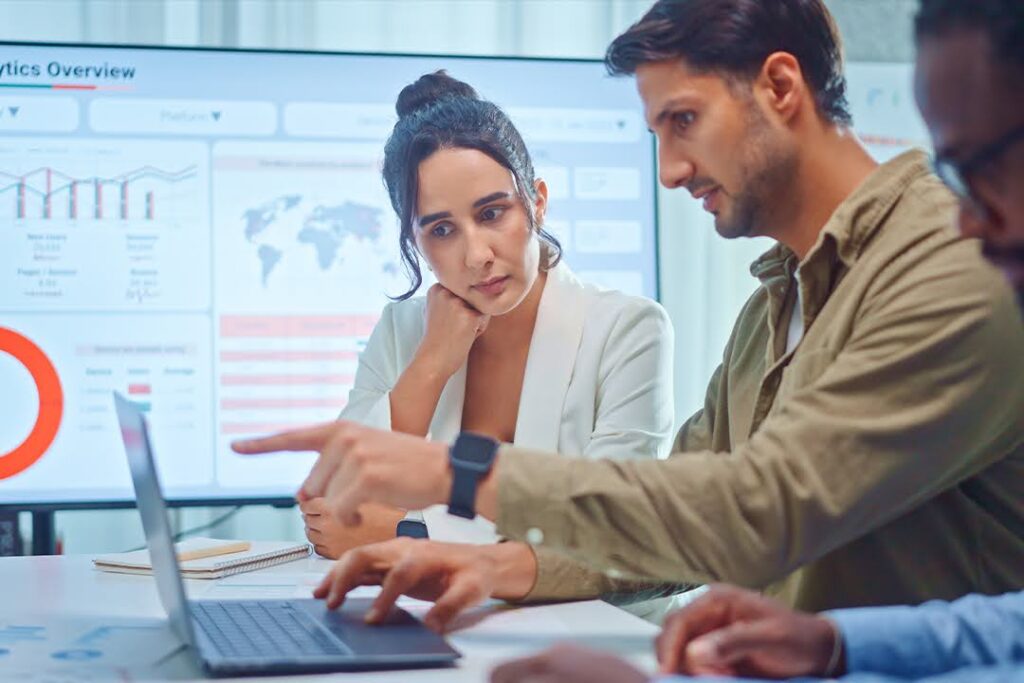 Two professionals review data on a laptop; charts and graphs are displayed on a large screen in the background.